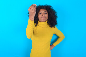 Young woman with afro hairstyle wearing yellow turtleneck over blue background angry gesturing typical italian gesture with hand, looking to camera