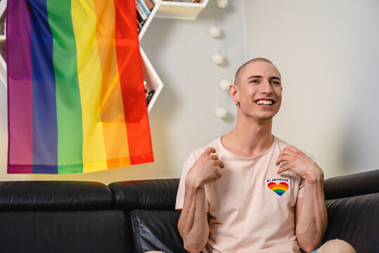 Bald queer caucasian activist sitting on a sofa, watching tv, and smiling with their hands up. Rainbow flag in the background. High quality photo