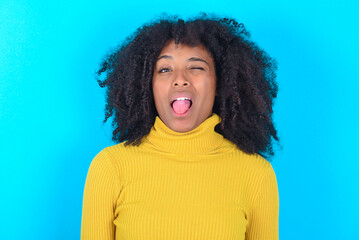 Young woman with afro hairstyle wearing yellow turtleneck over blue background sticking tongue out happy with funny expression. Emotion concept.