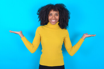 Cheerful cheery optimistic Young woman with afro hairstyle wearing yellow turtleneck over blue background holding two palms copy space