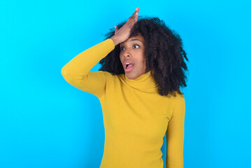 Young woman with afro hairstyle wearing yellow turtleneck over blue background surprised with hand...