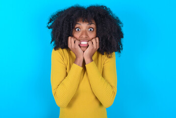 Fearful Young woman with afro hairstyle wearing yellow turtleneck over blue background keeps hands near mouth, feels frightened and scared,  has a phobia,  Shock and frighted concept.