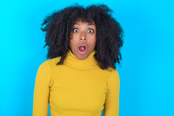 Oh my God. Surprised Young woman with afro hairstyle wearing yellow turtleneck over blue background stares at camera with shocked expression exclaims with unexpectedness,