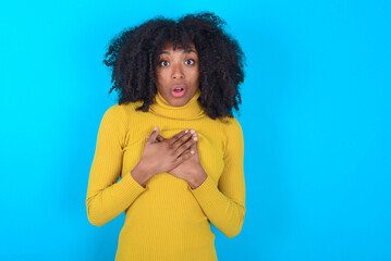 Scared Young woman with afro hairstyle wearing yellow turtleneck looks with frightened expression, keeps hands on chest, being puzzled to notice something strange, People, hush reaction and emotions.