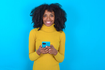 Young woman with afro hairstyle wearing yellow turtleneck over blue wall enjoys distant communication, uses mobile phone, surfs fast unlimited internet, has pleasant smile, makes shopping online,