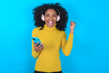 Positive Young woman with afro hairstyle wearing yellow turtleneck over blue background holds modern cell phone connected to headphones, clenches fist from good emotions, exclaims with joy,