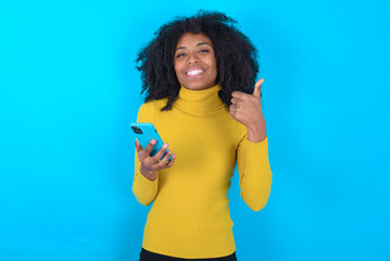 Portrait of Young woman with afro hairstyle wearing yellow turtleneck over blue background using and texting with smartphone  happy with big smile doing ok sign, thumb up with fingers, excellent sign