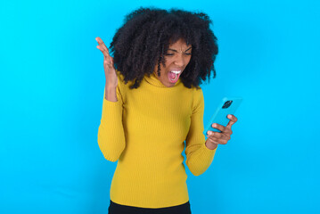 Angry Young woman with afro hairstyle wearing yellow turtleneck over blue background screaming on...