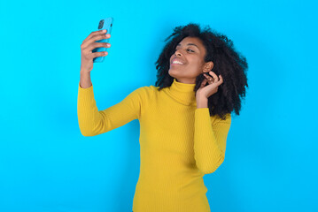 Young woman with afro hairstyle wearing yellow turtleneck over blue background smiling and taking a...