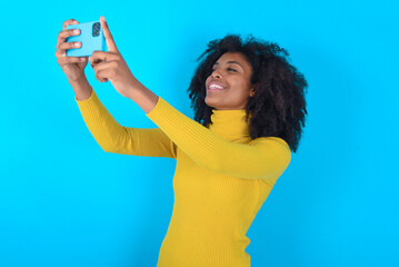 Young woman with afro hairstyle wearing yellow turtleneck over blue background taking a selfie to...