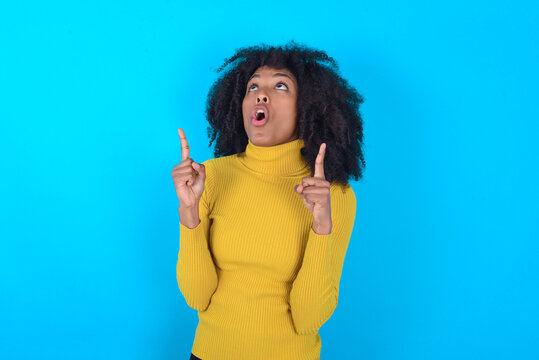 Young woman with afro hairstyle wearing yellow turtleneck over blue background amazed and surprised looking up and pointing with fingers and raised arms.