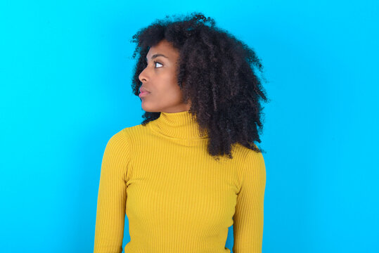 Close up side profile photo Young woman with afro hairstyle wearing yellow turtleneck over blue background not smiling attentive listen concentrated