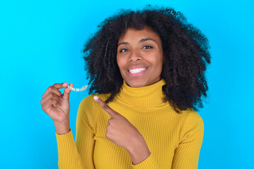 Young woman with afro hairstyle wearing yellow turtleneck over blue background holding an invisible aligner and pointing at it. Dental healthcare and confidence concept.
