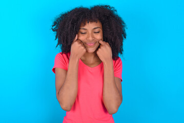 Pleased Young girl with afro hairstyle wearing pink t-shirt over blue background with closed eyes keeps hands near cheeks and smiles tenderly imagines something very pleasant