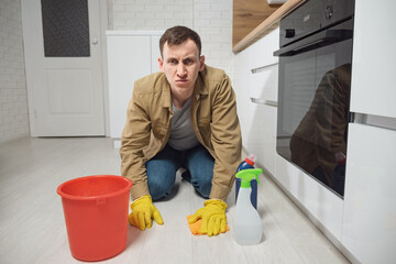 Man cleans laminate floor with detergent and water in plastic bucket smiling in modern kitchen. Young male housekeeper takes care of apartment doing chores, closeup