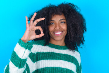 Young woman with afro hairstyle wearing striped sweater over blue background hold hand arm okey symbol toothy approve advising novelty news