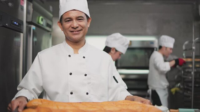 A senior Asian chef male  bakers in a white chef dresses uniform and hats standing cutting on a counter with many baked and unbaked bread on trays at a bakery kitchen restaurant.