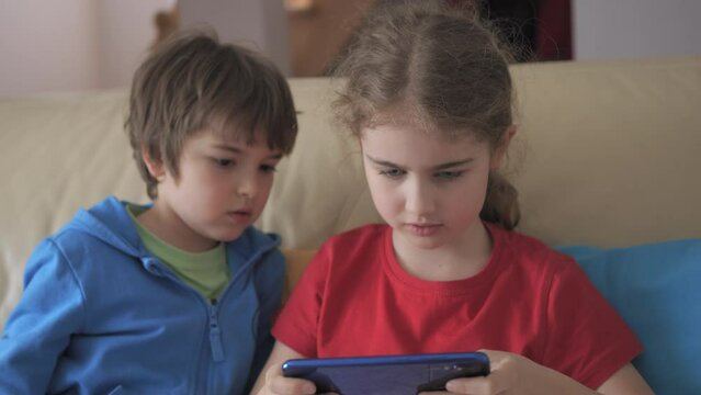 Children Playing Games In Phone at Home on Couch. Kids Playing Video Game on Mobile Phone. Boy and Girl Plays Video Game Smartphone on Sofa Friends Using Phone for Gaming Online Education Social Media