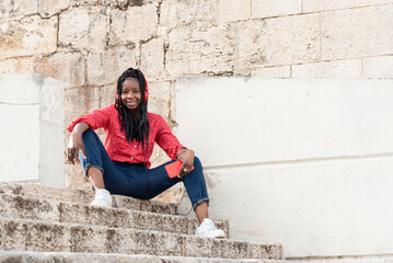 a smiling young african american woman with braids listening to music with red headphones sitting...