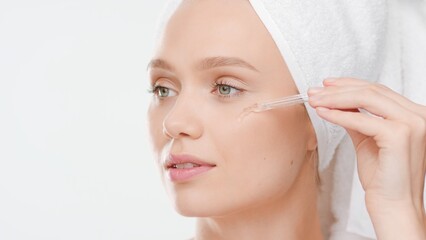 Big close-up shot of young Caucasian woman with hair-drying towel on her head applies face serum on her cheek from dropper on white background