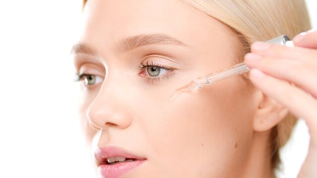 Big close-up shot of attractive European young woman applies face serum on her cheek from dropper on white background