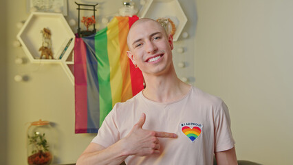 Happy caucasian gay man smiling at camera, pointing at a rainbow heart on his t-shirt, posing in his apartment, and standing in front of rainbow pride flag. High quality photo