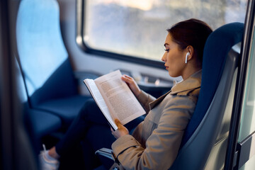 Young woman reads novel while listening music over earbuds in train.