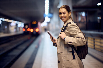 Happy woman uses cell phone while waiting at subway and looking at camera.
