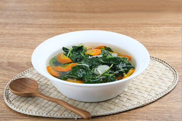 Sayur Bening Bayam, Spinach Clear Vegetable. Indonesian food of spinach, spinach soup with carrots. Served in white bowl on wooden background. 