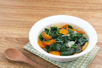 Sayur Bening Bayam, Spinach Clear Vegetable. Indonesian food of spinach, spinach soup with carrots. Served in white bowl on wooden background. 
