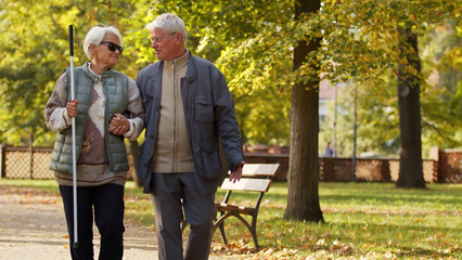 Senior Caucasian blind lady with glasses and a stick relies on her husband while having a walk in the park disabled blind people support concept medium full shot copy space. High quality photo