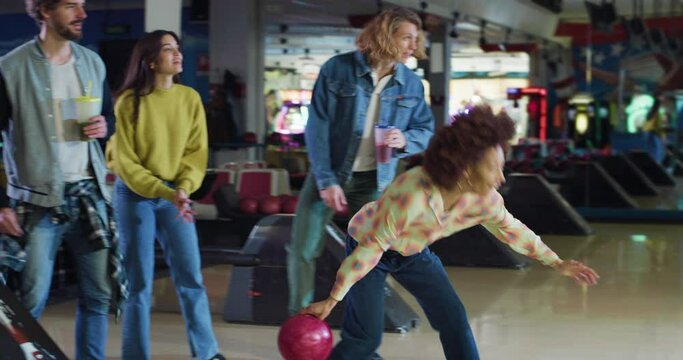 Cinematic shot of happy young african female bowler throwing strike ball and exulting victory while having fun to play bowling together with multiracial college friends on weekend in sporting club.