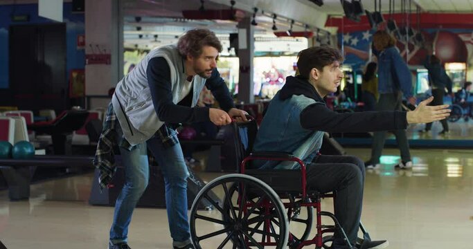 Cinematic shot of young man with disability who uses wheelchair throwing strike ball and exulting victory while having fun to play bowling together with his college friend on weekend in sporting club.