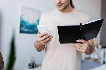 cropped view of man holding mobile phone and notebook at home.