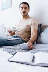 blurred man holding cup of coffee while reaching smartphone in bedroom.