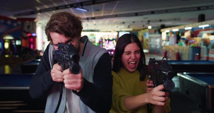Cinematic shot of young happy interracial couple shot with laser infrared guns while having fun to play shooting simulation game together on fairground in amusement park on weekend.