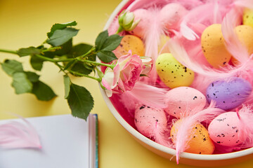 Festive Easter background. Pink bowl with colorful eggs, pink feathers, pink rose and notepad. Happy Easter concept. Post card on yellow background.