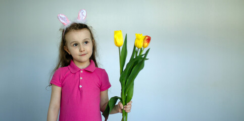 Obraz na płótnie Canvas A little girl with bunny ears smiles funny on a blue background with a bouquet of tulips in her hands. The Easter bunny is a symbol of the Bright Easter holiday. Postcard, copy space