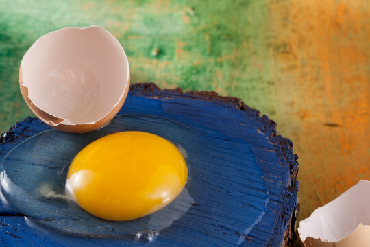 A broken raw chicken egg lying on a piece of cracked blue wood. The shell lies next to it. Blurred background in green and orange colors.