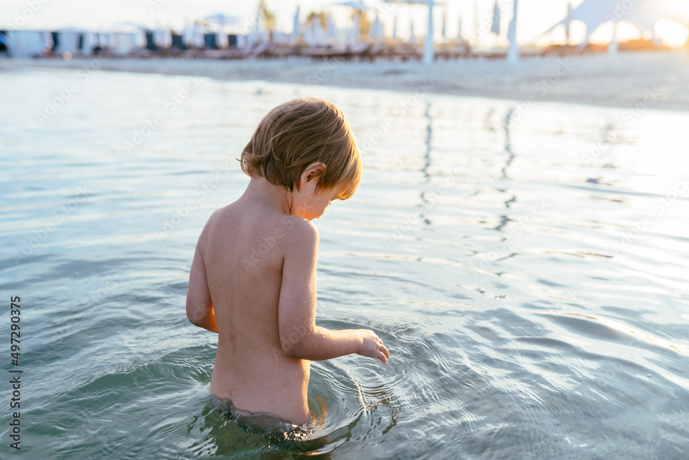 Wall mural cute adorable cheerful caucasian little blond boy stand carefully in the water at lake or river beac