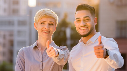 Multiethnic two business partners colleagues caucasian woman and hispanic man put thumbs up standing outdoors at sunset. Multiracial couple businessman and businesswoman posing in sunlight show like