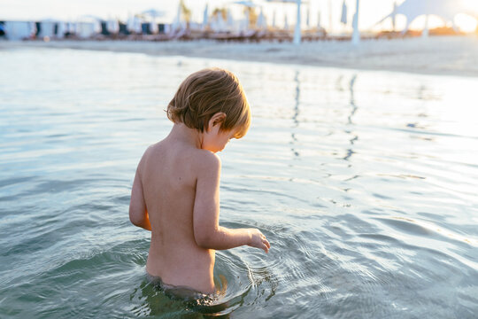 Cute adorable cheerful caucasian little blond boy stand carefully in the water at lake or river beach water on warm sunset evening time outdoors. Happy childhood vacation at countryside concept. - Powered by Adobe