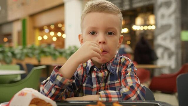 Caucasian boy satisfies his hunger by eating potatoes in a rustic way from fast food. Portrait hungry blond 4-year-old boy eating fast food, fried Idaho potatoes.
