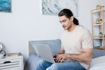 freelancer with long hair sitting on bed and using laptop.
