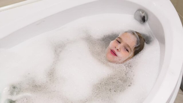 a little cheerful girl washes and plays in the bath with foam