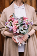 Very nice young woman holding small and beautiful bouquet of fresh eustoma, matthiola, roses, eucalyptus, chamellacium in purple and pink colors, bouquet close up on the wooden background - 497292764