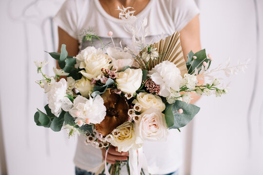 Very nice young woman holding big and beautiful bouquet of fresh roses, carnations, chamellacium, eucalyptus and dry palm leaves in white and brown colors, cropped photo, bouquet close up