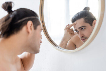 shirtless man with hair bun on head applying face cream and looking at mirror.