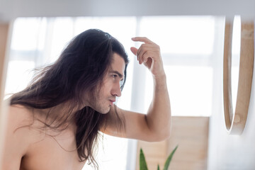 side view of shirtless man styling long hair near mirror.