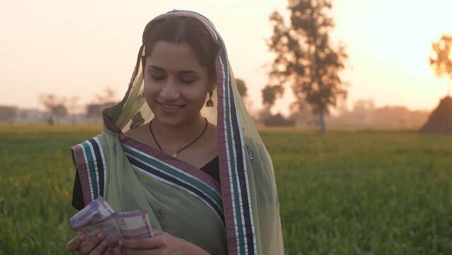 A female farmer happily counting her hard-earned money in her farmland - monthly income  future investment  agricultural loan. A pretty village lady holding a bundle of Indian currency notes - Fina...